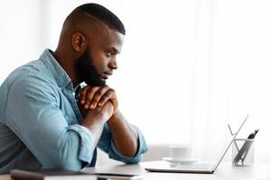 Thoughtful Black Writer Working With Laptop At Home Office. African American Freelancer Man Sitting At Desk And Looking At Computer Screen With Pensive Face Expression, Side View With Copy Space photo