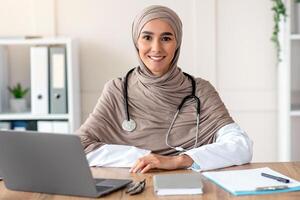 Portrait of smiling female doctor young beautiful woman in hijab with stethoscope posing in her cabinet, sitting at workdesk with medical chart, notebook and laptop in clinic, copy space photo