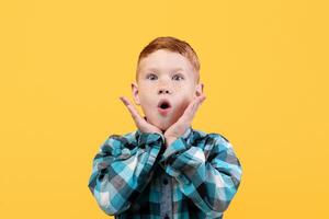 Surprised little boy in casual shirt, face expression concept. Shocked redhead kid keeping mouth open and touching his face, seeing something shocking, yellow studio background, copy space. photo