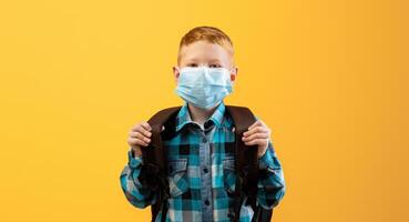 Schoolboy with backpack wearing protective face mask over yellow studio background. Redhead kid schooler using protection against coronavirus while going to school during COVID-19 pandemic photo