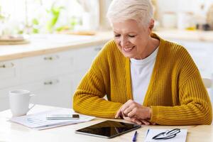 Joyful grey-haired senior woman using digital tablet in kitchen, taking notes, counting month spendings, copy space. Smiling old lady grandmother enjoying nice mobile application photo