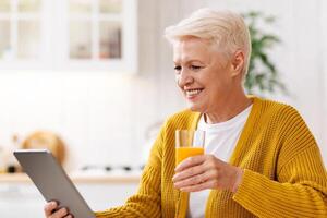 Joyful old grey-haired woman eating healthy food and using digital tablet in kitchen, empty space. Happy grandmother with glass of fresh juice reading culinary blog on modern pad photo