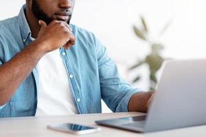Busy Black Entrepreneur Working With Laptop At Office, Unrecognizable African Businessman Sitting At Desk, Looking At Computer Screen And Touching Chin, Closeup Shot, Cropped Image photo