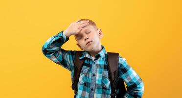 Exhausted kid schooler with heavy backpack touching his forehead on yellow studio background. Tired redhead boy in plaids shirt got tired of studying, suffering from burnout in school photo