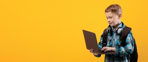 Technologies in school education. Smart schoolboy with backpack and headset holding laptop over yellow studio background, copy space. Cute kid using laptop for studying, doing homework, websurfing photo