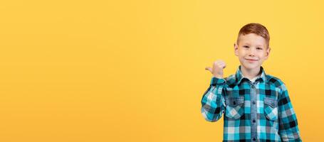 Smiling red-haired caucasian boy aiming at free space behind his back, empty space for text or advertisement, panorama. Cheerful ginger kid pointing with thumb at copy space over yellow background photo