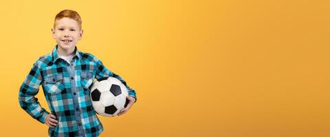 Active cheerful ginger boy posing with soccer ball on yellow studio background, copy space. Happy kid playing football, holding ball. Kids and teens sport lifestyle concept photo