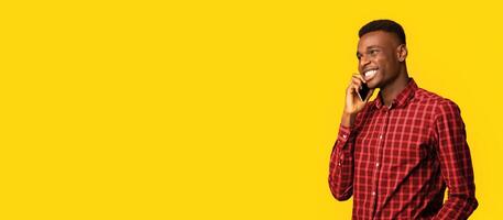 Mobile Communication. Positive African American Guy Talking On Cellphone And Looking Away, Young Black Man Enjoying Pleasant Conversation While Standing Isolated Over Yellow Background, Copy Space photo