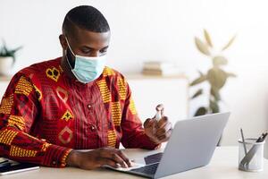 Hygiene On Workplace. African Man In Medical Mask Applying Disinfectant Spray On Laptop Keyboard For Coronavirus Prevention, Black Man In Traditional Clothes Sanitizing Computer Surface, Copy Space photo