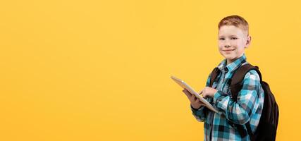 Cheerful schoolboy using digital tablet for education, doing homework on yellow studio background, copy space. Smiling redhead boy with backpack holding pad, websurfing or using new application photo