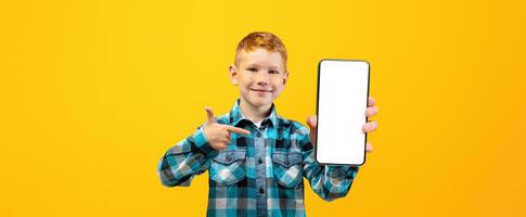 Smiling ginger boy in casual shirt pointing at modern mobile phone with empty screen over yellow studio background, mockup. Happy schoolboy with smartphone for education or entertainment, copy space photo