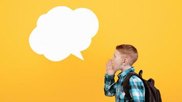Side view of ginger schoolboy with backpack saying something loud and empty communication cloud over yellow studio background, copy space. Boy with schoolbag looking for attention photo