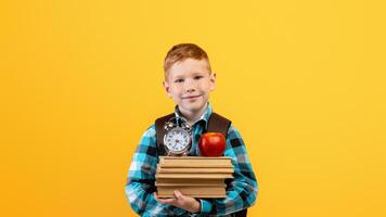 Cheerful ginger kid schooler with backpack holding heap of books, red apple and watch on yellow studio background, copy space. Schedule, timing for children schoolers concept photo