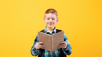 Joyful redhead schoolboy in shirt reading book, yellow studio background. Happy kid with backpack enjoying exciting book, love studying, copy space. School educational system concept photo