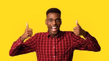 All Is Great. Cheerful african american guy showing thumbs up, making approval gesture with both hands and smiling at camera, standing isolated on yellow studio background, closeup with free space photo