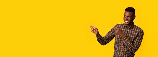 Look There. Cheerful African American Guy Pointing Aside At Copy Space Over Yellow Background With Two Fingers, Positive Black Male Showing Free Place For Your Design Or Advertisement, Panorama photo