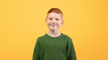 Portrait of happy cute ginger boy in knitted green sweater posing on yellow studio background, copy space. Redhead kid in casual outfit smiling at camera, having good mood. Childhood concept photo