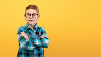 Intelligent ginger boy in glasses standing with arms crossed on chest, posing over yellow studio background. Cute smart red-haired kid nerd in plaids shirt smiling at camera, copy space photo