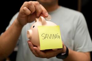 Man Depositing Coin into Piggy Bank with Saving Note. A man's hand is shown actively depositing a coin into a pink piggy bank. financial planning, making deposits, and setting financial goals. photo