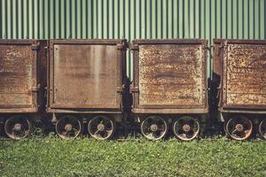 Old rusted train cars lined against a green corrugated backdrop in an abandoned area photo
