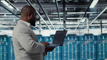 Server room infrastructure engineer scripting automated workflows using software on laptop. Data center programmer running predictive maintenance scans using notebook, camera B video