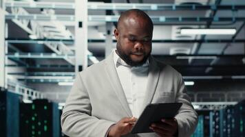 Close up of technician walking in data center, using tablet to check equipment for system stability. IT expert overseeing server farm, using device to configure rigs, optimizing performance, camera B video