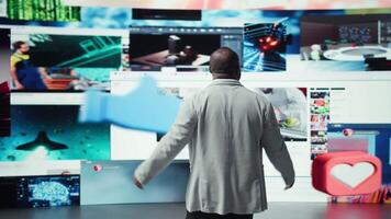 Cheerful user enjoying unlimited entertainment in modern digital environment. Happy man stands with open arms in front of giant digital wall streaming online videos and social media content, camera A