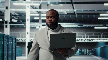 Technician walking in data center, configuring storage devices for minimal latency in AI workloads. African american employee using device in server farm to build neural networks, camera B video