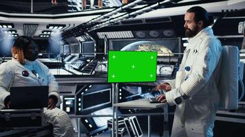 Astronaut monitoring system diagnostics on green screen display, talking to colleague during space mission. Teamworking cosmonauts using chroma key monitor during mankind survival expedition, camera A video