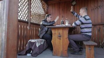 Two men sit at the table on terrace and drink beer from plastic bottles. Happy and satisfied. video