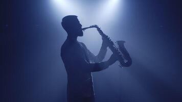 Backlit silhouette of saxophonist man with saxophone standing in dark nightclub disco studio and start playing sax. Light appears and illuminates the man. Isolated on black background. Slow motion video