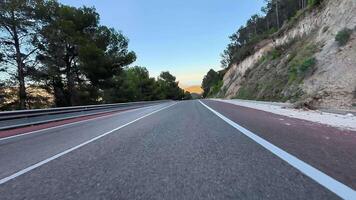 The vibrant sunset skies beautifully stretch over a winding road that is bordered by lush trees and rocky hills photo