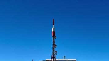 A striking red and white telecom tower prominently stands out on a lush green hillside beneath a bright clear blue sky photo