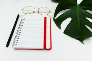 Open red notebook with pencil, glasses and monstera leaf. School notebook on white background, spiral notepad on table photo