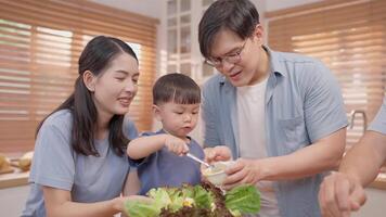 A family enjoys quality time in a bright kitchen, with a parent guiding a child on how to prepare a healthy dish using fresh ingredients like lettuce. They share smiles and laughter while cooking. video