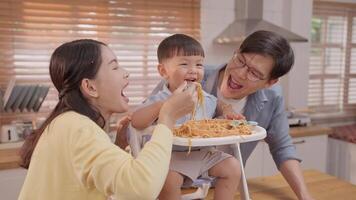 A family bonds while enjoying a spaghetti meal at home. The toddler happily feeds himself, while parents engage joyfully, creating a warm atmosphere in their kitchen. video