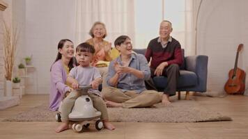 A joyful family gathers in a warm living room. A toddler happily rides a toy, while family members, including grandparents and parents, share smiles and laughter in a vibrant atmosphere. video