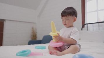 A young child sits on a bed, engaged in playing with vibrant stacking rings. The atmosphere is cheerful and sunny, enhancing the playful experience of exploration and learning. video