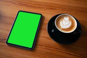 Top view of modern digital device with blank green screen and cappuccino for work or relaxation themes. photo