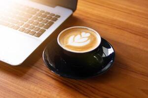 Side shot of latte art in black cup with laptop keyboard in soft background light. photo