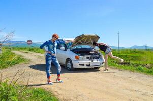 Red-haired woman with a screwdriver and pliers on high heels standing near a broken car with open hood on a country road and barrier with stop sign photo
