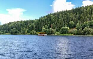 View from the river to a small pier on the other bank, with a coniferous forest in the background, on a summer day photo