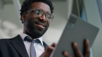fechar acima africano americano feliz sorridente o negócio homem dentro escritório usando gráficos digital tábua computador dispositivo sorrir despreocupado étnico masculino homem de negocios cara trabalhador navegando tela sensível ao toque conectados trabalhos Internet video