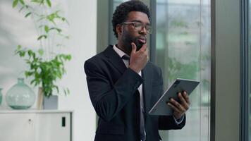 African American pensive thinking business man in office looking gazing out window using digital tablet computer device smiling thoughtful businessman guy creative worker find idea browsing screen video