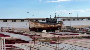 An old, rusted fishing boat is docked in a desolate shipyard, all under a bright blue sky that adds to the scene photo
