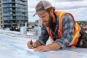 A construction worker in safety gear is carefully marking construction plans on a rooftop in a busy urban environment during daylight photo