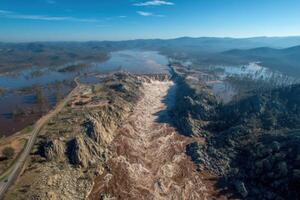 Aerial view highlights a rushing water flow and extensive flooding in a rugged landscape with hills and trees under a clear blue sky photo