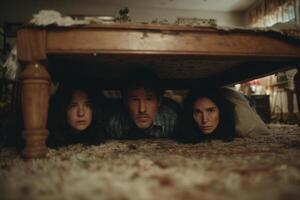 A man and two women crouch together under a table in a dark living room, showcasing a tense atmosphere photo