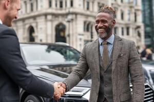 Two men in formal attire greet each other with a handshake while standing near an elegant car outside a modern building photo