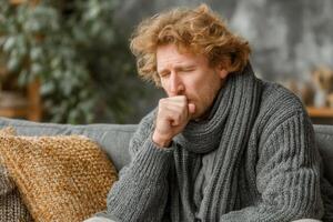A man is seated comfortably on a couch, coughing softly while bundled up in a cozy sweater and scarf indoors, suggesting he feels unwell photo